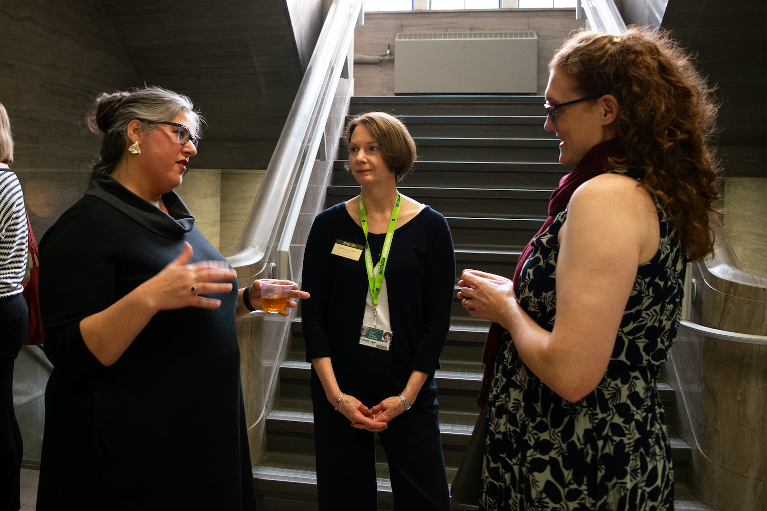 Three people converse near a stairway during an indoor university event or reception.