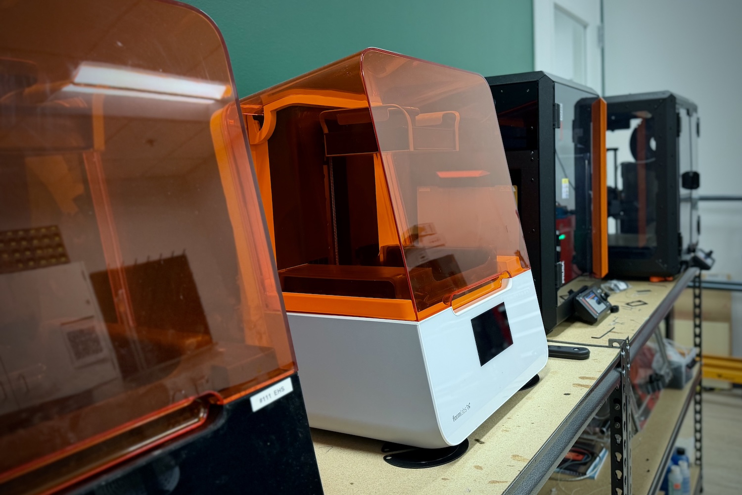 Close-up view of multiple resin 3D printers on a workbench, showing orange-tinted protective covers, white and black printer bodies, and fabrication equipment arranged along a lab shelf.