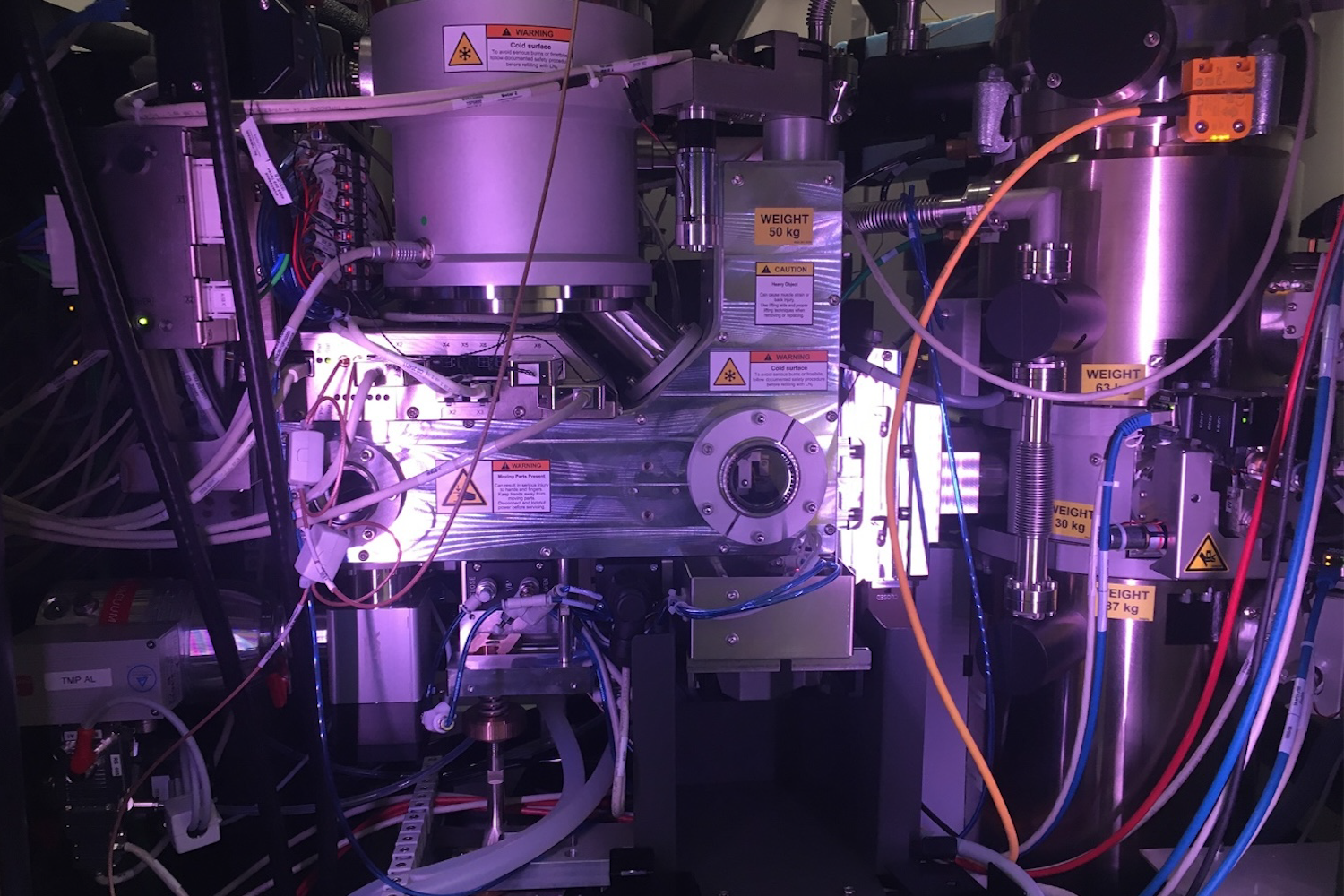 Close-up view inside a cryo-electron microscope showing metallic instrument components, vacuum chambers, warning labels, cables, tubing, and purple internal illumination.