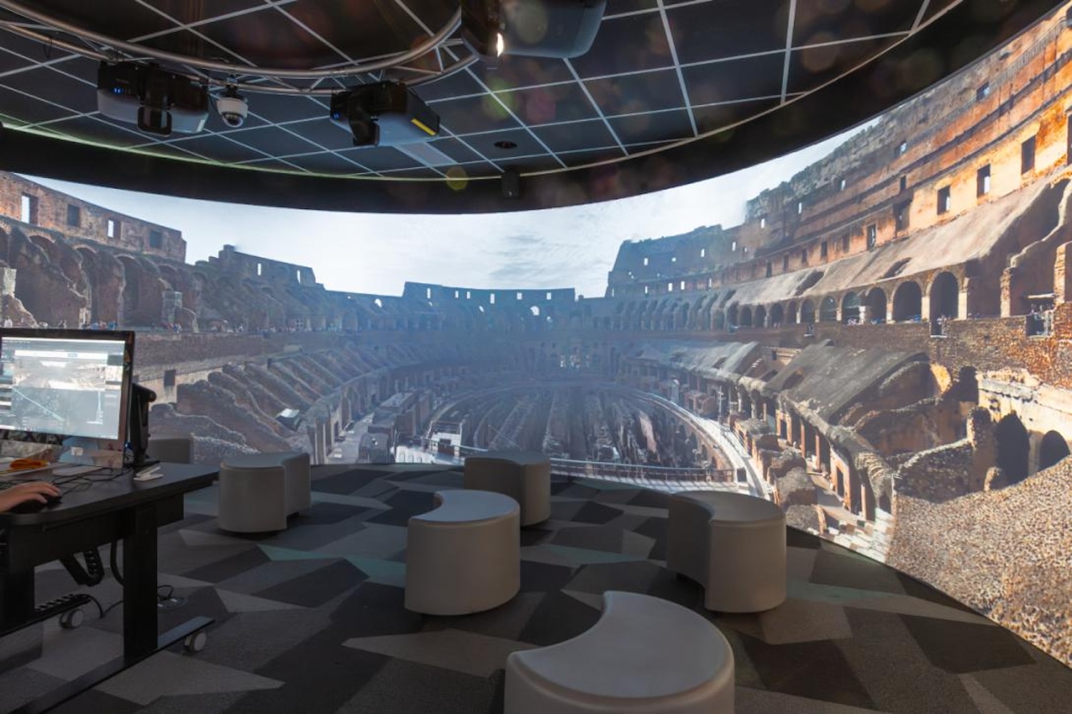 Immersive 360-degree visualization room with curved wall projection showing the Roman Colosseum, ceiling-mounted projectors, computer workstation, and mod