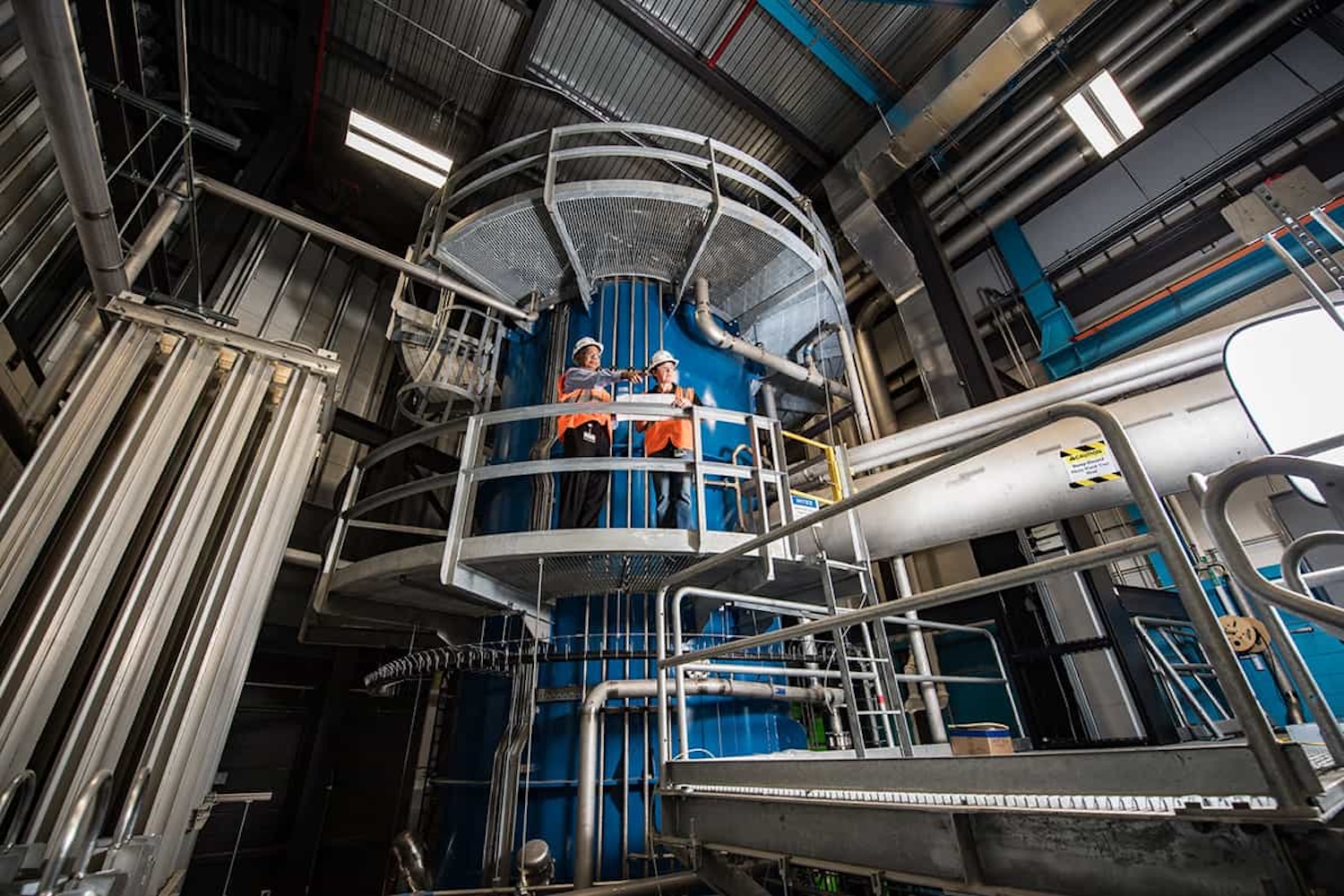 Interior of the Facility for Rare Isotope Beams at Michigan State University, showing workers on an elevated platform surrounded by large blue accelerator equipment, metal railings, pipes, cables, and industrial infrastructure.