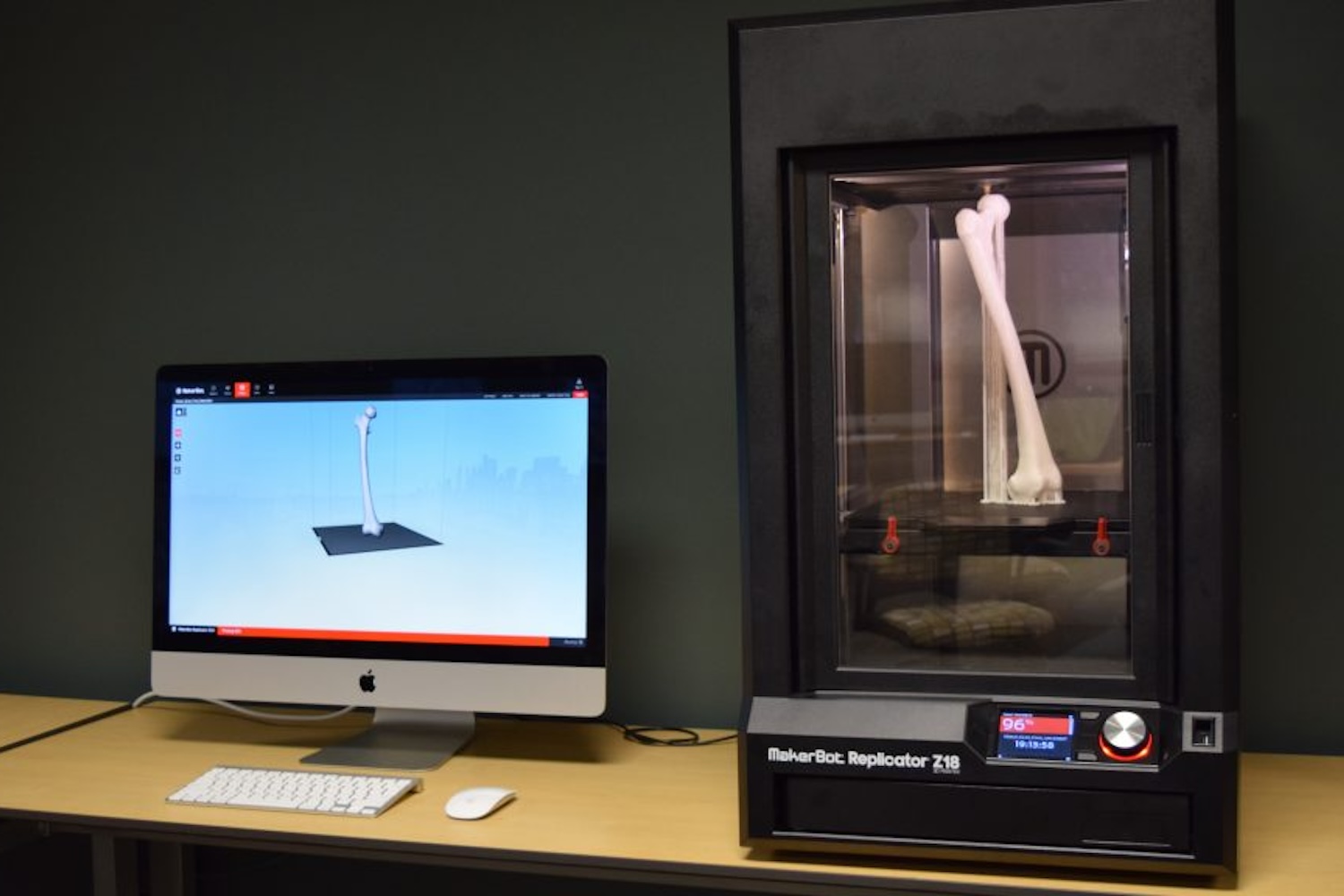 3D printer creating a bone-shaped model beside a computer displaying the corresponding digital design.