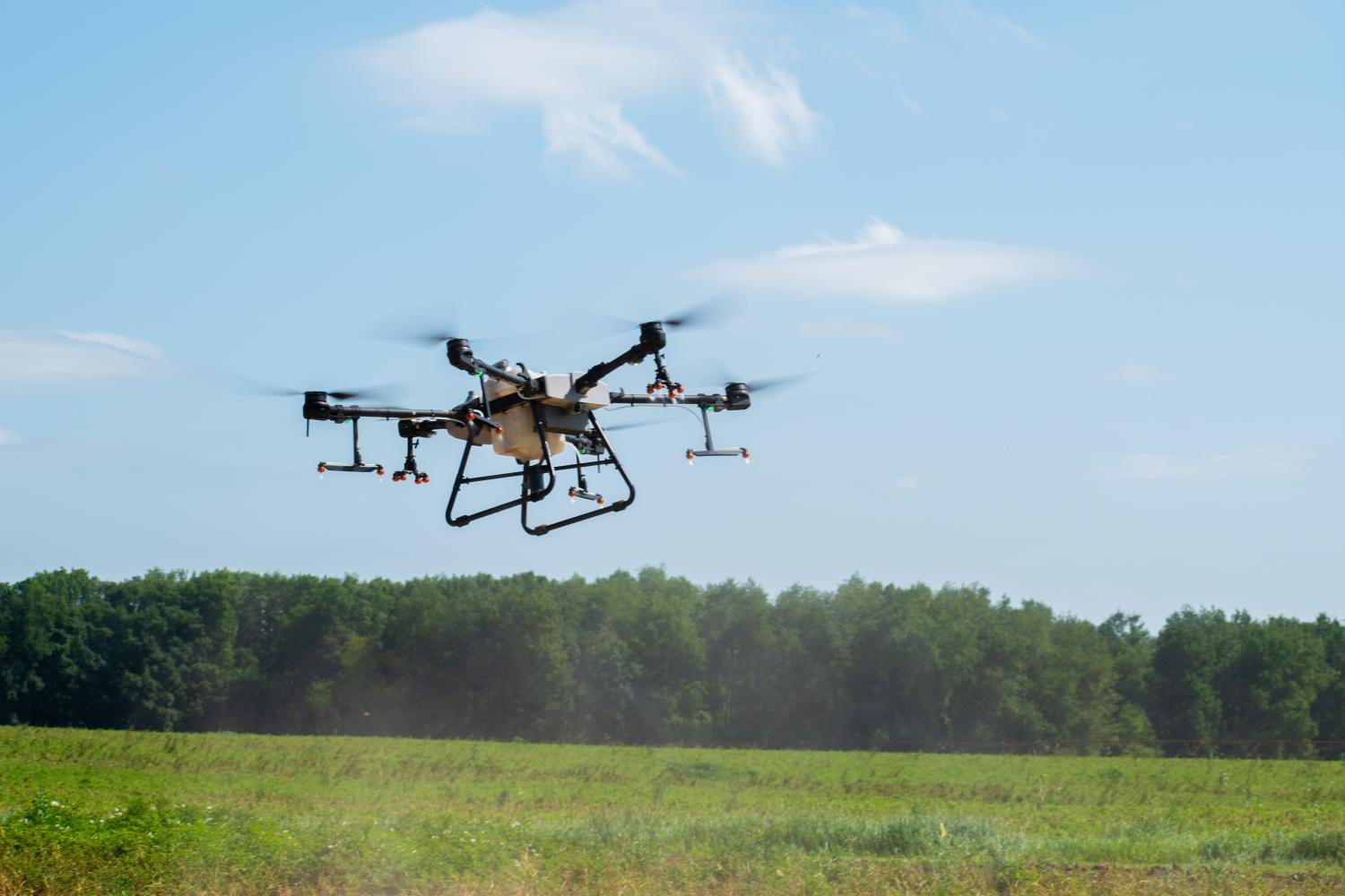 Large agricultural drone flying over a green field with trees in the background under a blue sky, with blurred rotor motion and spray mist visible below the aircraft.