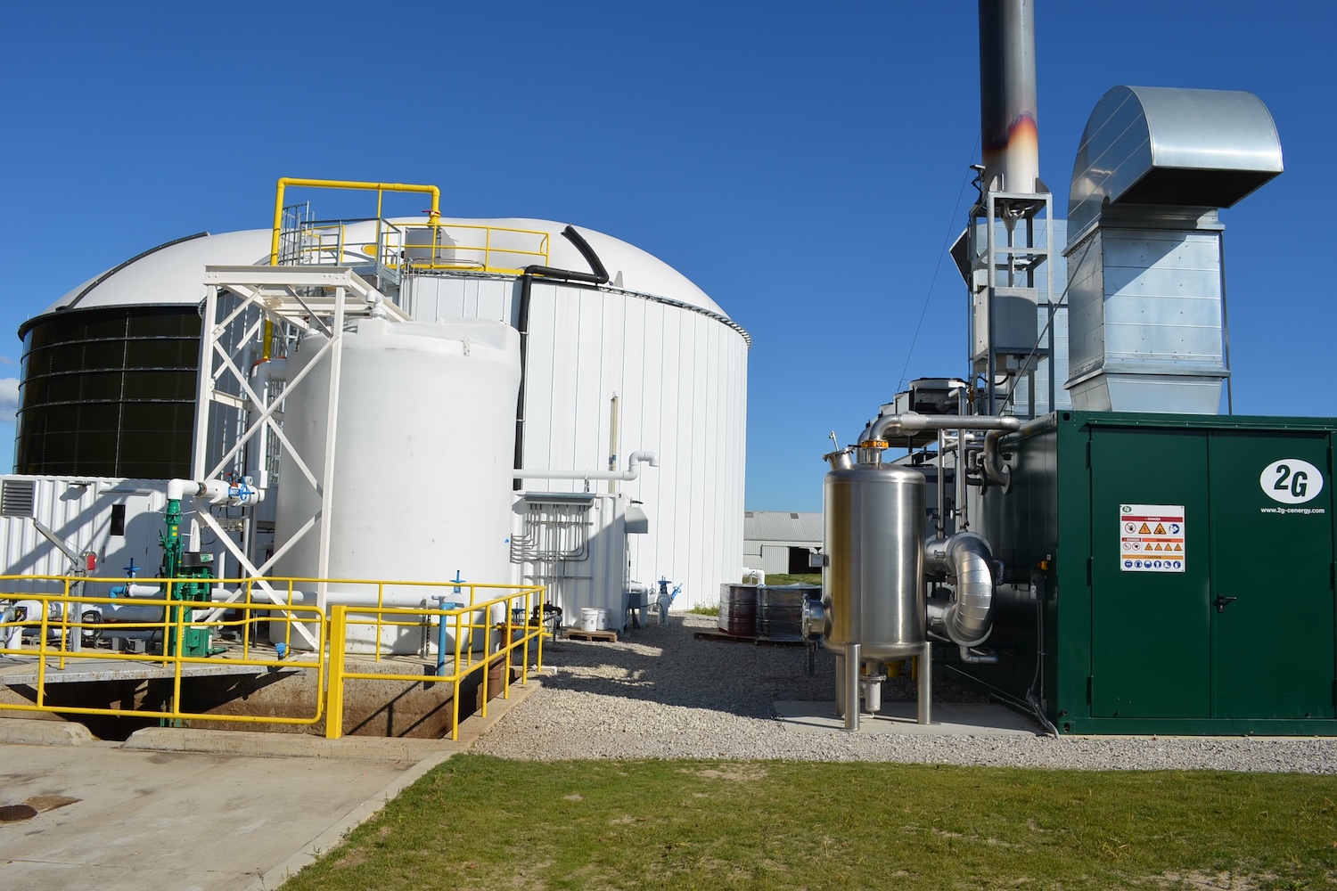 Anaerobic digester facility with large storage tanks, interconnected pipes, yellow safety railings, ventilation equipment, and a flare stack burning against a clear blue sky.