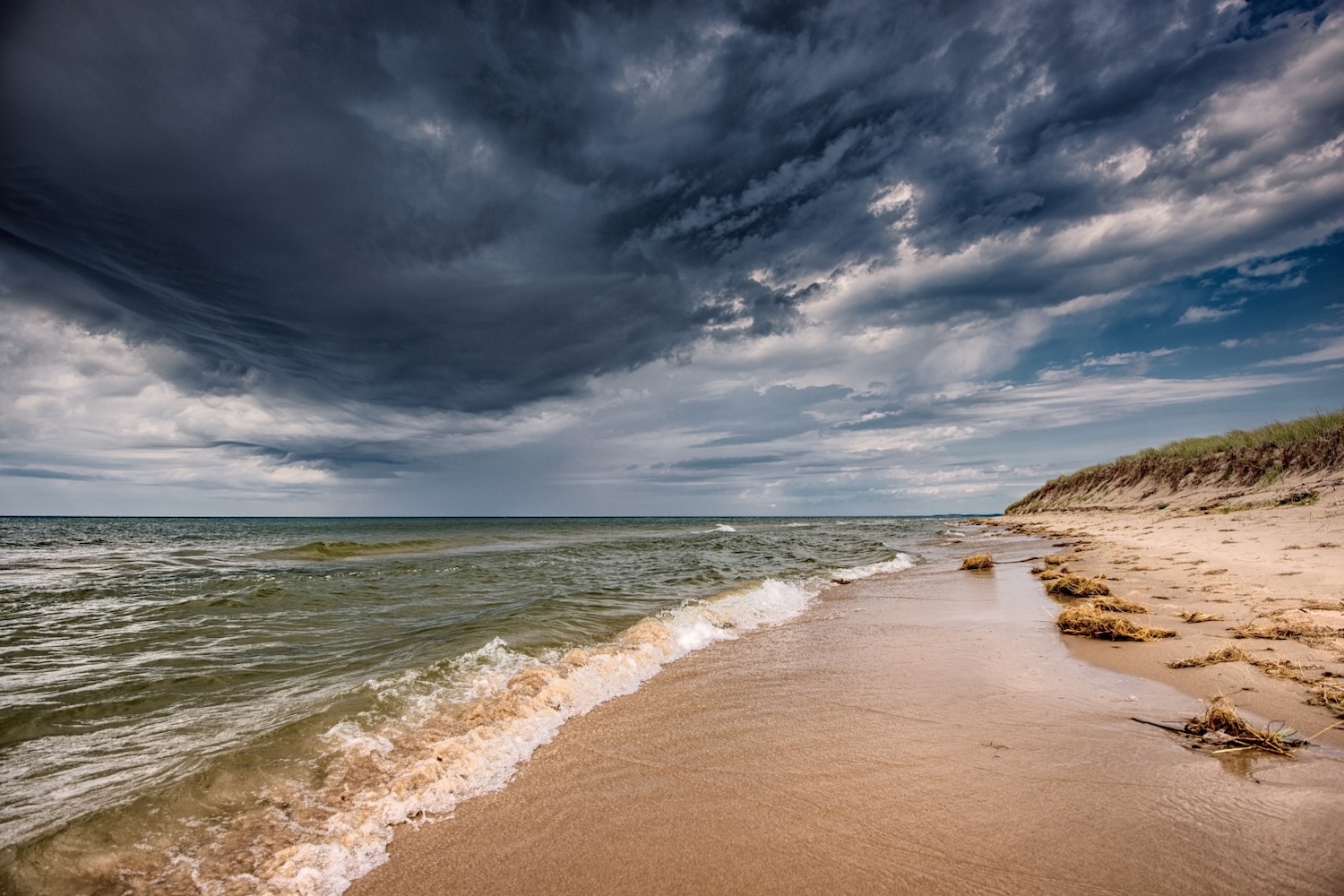 Sandy Lake Michigan shoreline at Nordhouse Dunes near Ludington State Park and Manistee National Forest, with waves washing onto the beach, dune grass along the shore, and dramatic dark storm clouds overhead.