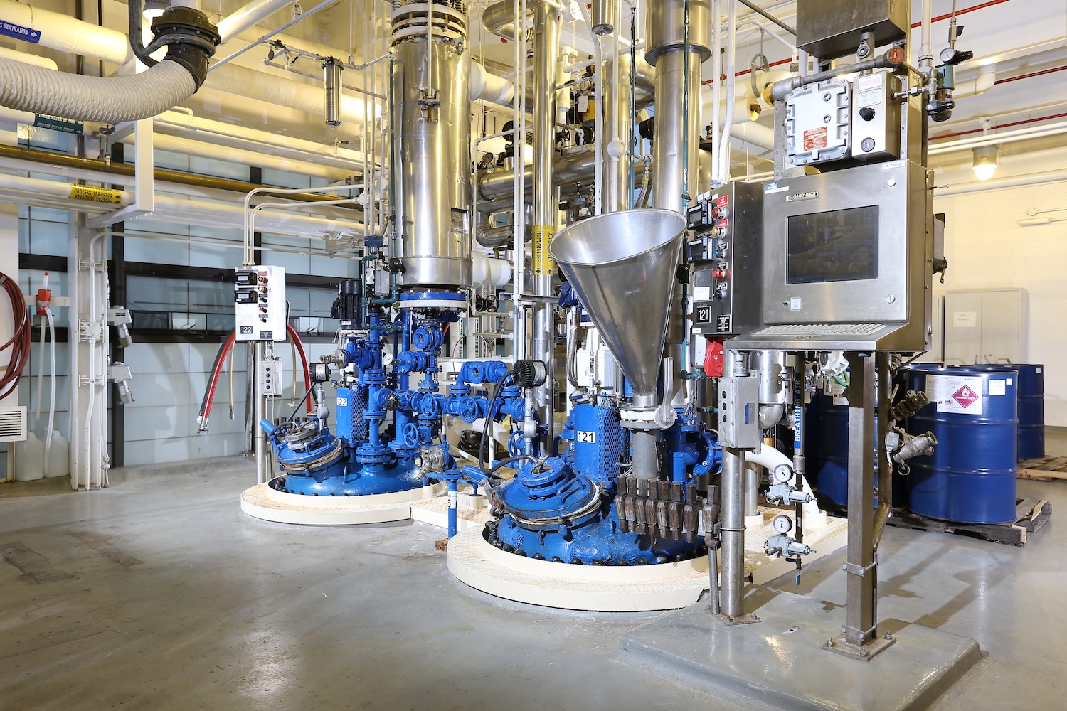 Pilot-scale chemical processing equipment inside an industrial chemical plant, including stainless steel vessels, blue pumps and valves, overhead piping, control panels, and chemical storage drums in a clean production area.