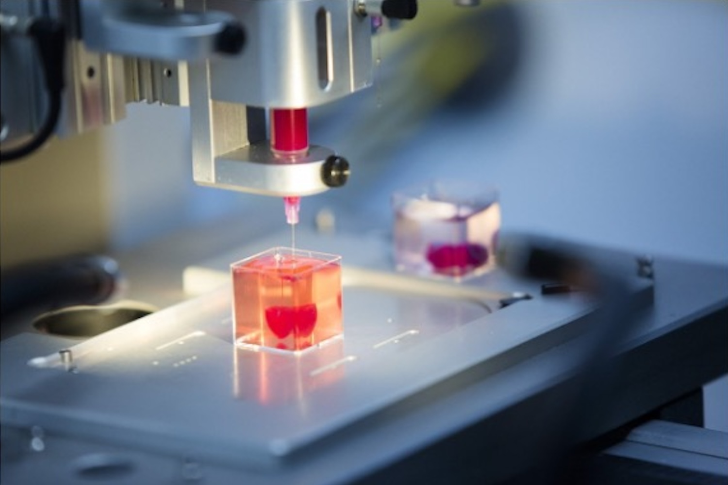 Close-up of a 3D printer or bioprinting instrument depositing red material into a clear sample container in a laboratory setting.