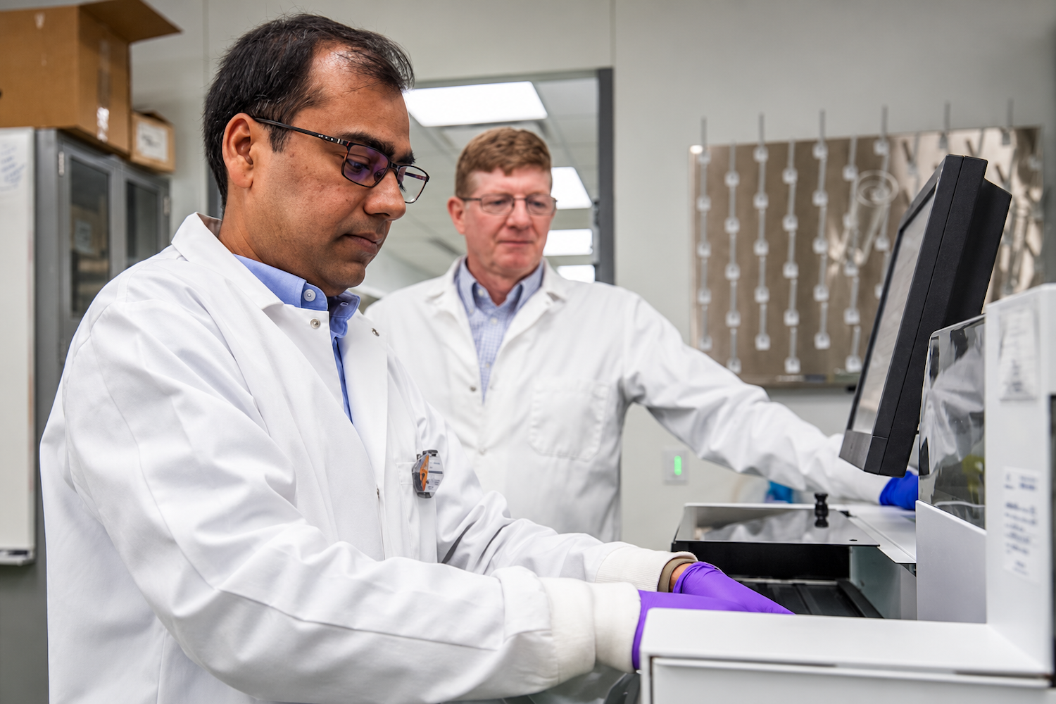 Two researchers in lab coats operate and observe laboratory instrumentation in a research facility.