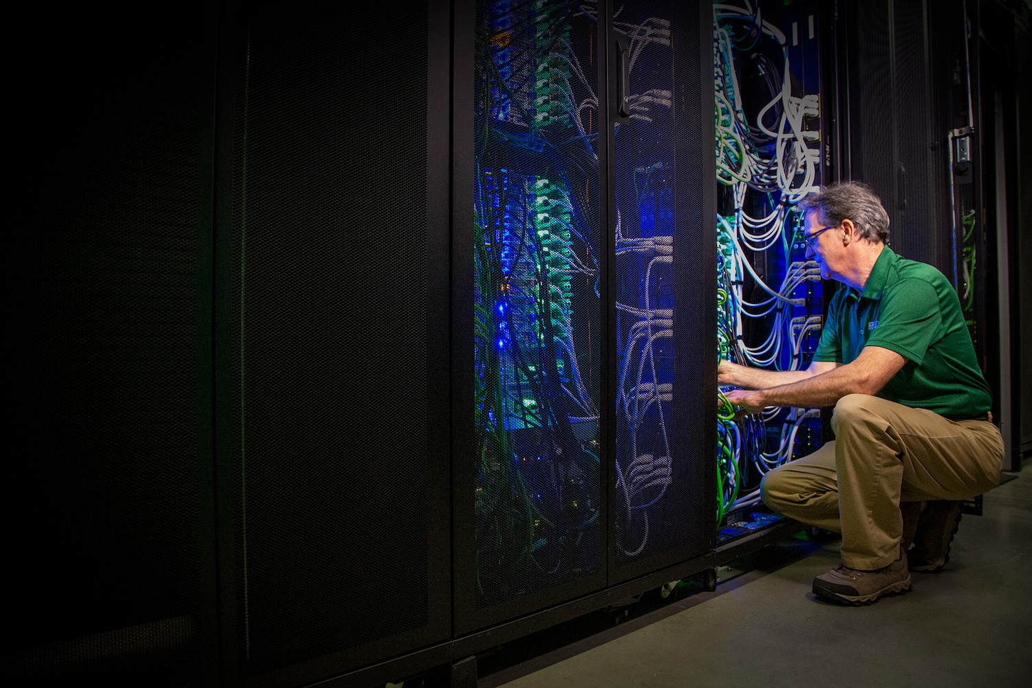 staff member working inside a data center server rack with network cables, illuminated server equipment, and dark server cabinets.